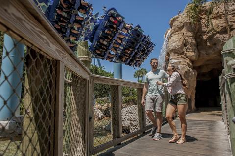 A couple walks across a wooden bridge at SeaWorld as a roller coaster full of riders swoops overhead.