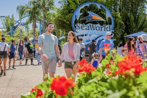 Couple holding hands and smiling while walking through SeaWorld Orlando, surrounded by other visitors and colorful flowers near the park entrance.