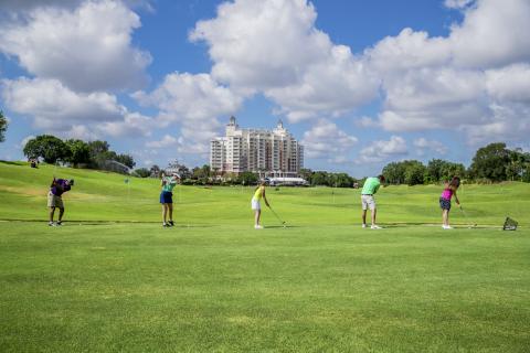 A group of five people practice their swings on a green driving range at Reunion Resort, with a tall resort building and a bright blue sky with scattered clouds in the background.