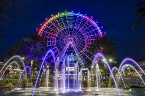 Nighttime view of The Wheel at ICON Park in Orlando, glowing with rainbow lights above a fountain display.