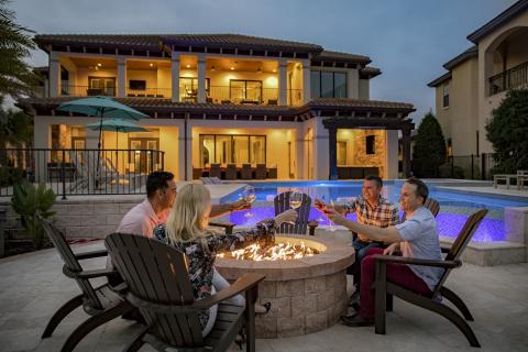 Friends gathered around a glowing fire pit at a luxury Kissimmee vacation home, toasting drinks beside the illuminated pool at dusk.
