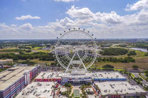 Aerial view of the Orlando Observation Wheel at ICON Park, surrounded by shops, restaurants, parking garages, and wide stretches of green landscape under a bright, partly cloudy sky.
