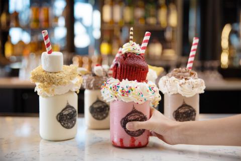Hand holding a pink milkshake in the foreground with three other milkshakes behind.