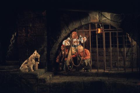 Dark scene from the Pirates of the Caribbean ride with 2 people behind bars and dog standing outside.