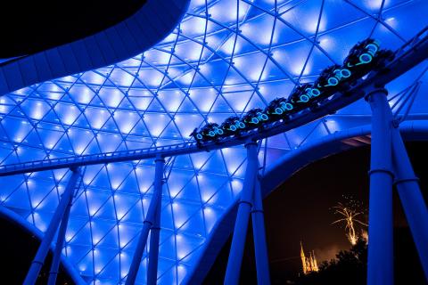 Blue and white lights glow while a roller coaster speeds below. 