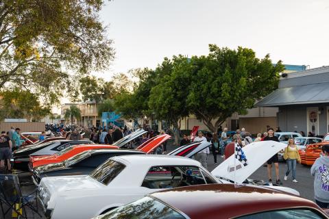 Classic cars with open hoods line a bustling street at a car show in Kissimmee, as visitors walk between the rows and admire the vehicles under evening light.
