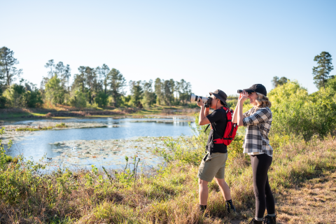 Two people use binoculars and a camera to observe wildlife beside a lakeside trail in Kissimmee.