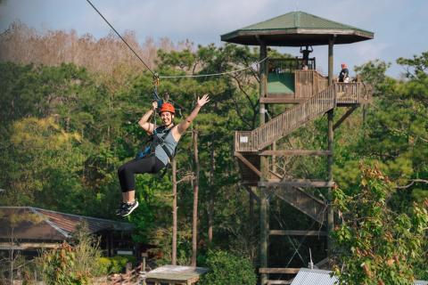 Woman smiling and waving while ziplining over treetops at Gatorland, near Kissimmee, Florida, on a sunny day.