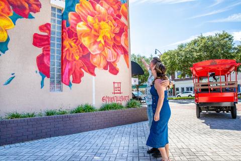 A couple looks at a large floral mural in downtown Kissimmee while standing near a red tour vehicle.