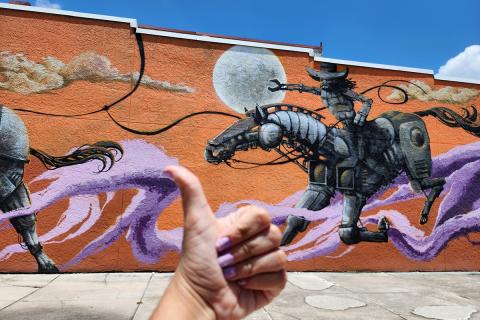 A person giving a thumbs-up in front of a vibrant Kissimmee mural featuring a futuristic robotic cowboy on horseback.