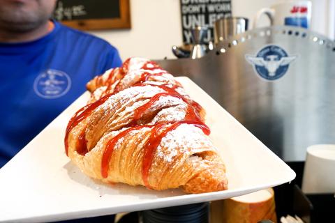 A powdered sugar–dusted croissant drizzled with strawberry sauce is served on a white plate inside a cozy café, with espresso equipment in the background.