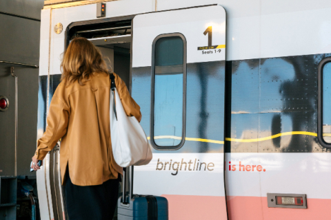 A traveler with a rolling suitcase boards a Brightline train at the station, walking toward the open door.