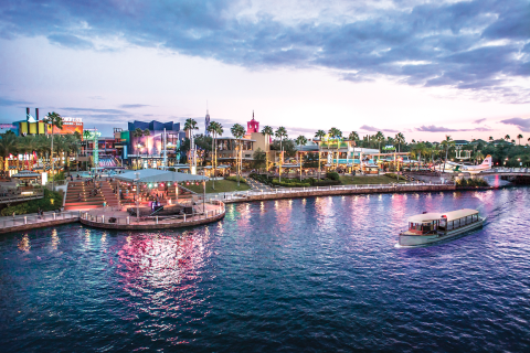 Evening view of Universal CityWalk in Orlando, Florida, with colorful lights reflecting on the water and a boat cruising along the canal near restaurants and entertainment venues.