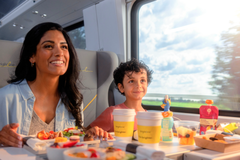 A mother and her young son enjoy a meal together while riding the Brightline train, with sunlight streaming through the window and a scenic view outside.