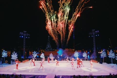 Fireworks explode above an outdoor ice-skating stage where performers in festive red-and-white costumes dance amid illuminated holiday trees at night.