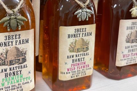 Bottles of raw natural honey from 3Beez Honey Farm in St. Cloud, Florida, displayed on a shelf with decorative bee charms tied around the tops.