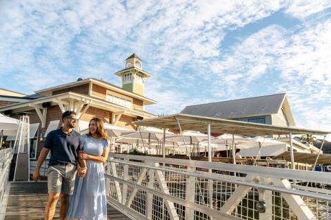 A couple walks hand in hand along the dock outside The BOATHOUSE at Disney Springs, smiling under a bright, partly cloudy sky with the restaurant’s tower and outdoor umbrellas in the background.