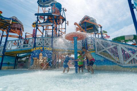 Father with two children getting splashed under a mushroom-shaped water feature at Island H2O Water Park in Kissimmee, Florida, with water slides and a “beach time” sign visible in the background.