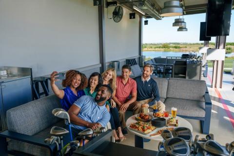 A group of young professionals sit on an outdoor couch closely to take a selfie. In front of them is a variety of small bites and surrounded by golf clubs.