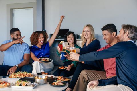 A group of adults sits together sharing food and drinks, laughing and raising their glasses in a celebratory toast.