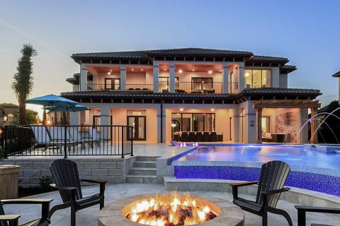 Luxury vacation home in Kissimmee featuring a spacious patio, infinity pool with fountains, and a cozy fire pit surrounded by lounge chairs at sunset.