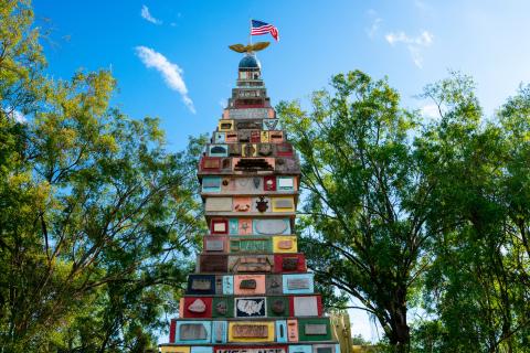 The upper half of the monument is pictured on a back drop with a blue sky and lots of oak trees. The front of the four sided, tapered monument shows many colored squares and rectangles with plaques ranging in size and shape. The very top of the monument is a small globe with gold wings and the American flag.
