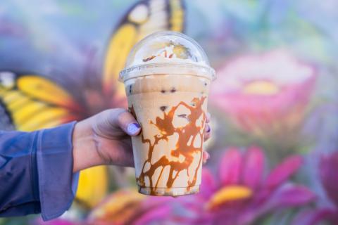 A person holds a caramel iced coffee against a mural of a Monarch butterfly landing on pink flowers. The person is wearing a blue, long sleeve button up shirt and has purple painted nails. The beverage has caramel drizzled on the side of the cup and is topped with whipped cream.