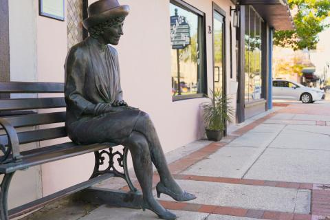 A bronze statue of Bette Sprinkle. She is sitting on a bronze bench positioned on a sidewalk with her legs crossed and smiling. She is wearing a hat, women's suit with a skirt, and kitten heels.
