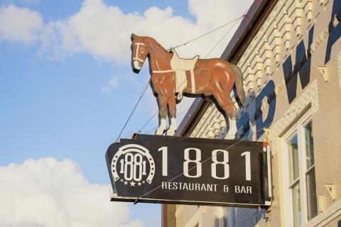 A brown horse with white hooves, ankles, and nose wears a saddle and is positioned above a black metal sign that reads 1881 Restaurant. The horse and sign are fixed to a beige-painted brick building with the blue cloudy sky as the backdrop.