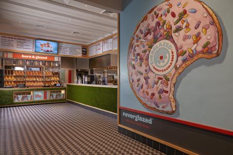 A detail of the decor shows a large pink doughnut with sprinkles on a light blue wall. The remainder of the store is just as colorful with many colors, including those of the doughnuts and images on the menu screen.