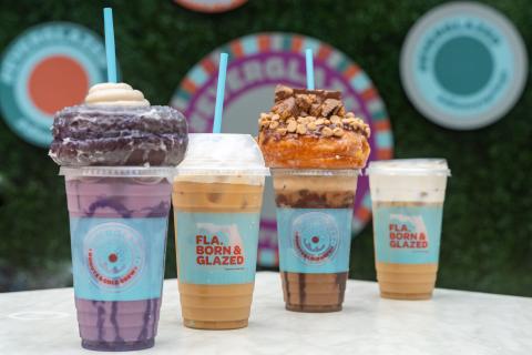 Four drinks are on a white tabletop with greenery in the background. The drink in the front is ube flavored, and it is light purple with dark purple swirls. It is topped with a glazed purple ube doughnut and white frosting. The drink next to it is a conventional cold brew with milk and topped with whipped cream. The next drink is peanut butter flavored and is light brown with chocolate swirls. It is topped with a glazed doughtnut with peanut butter cup crumbles. The last drink is another conventional brew.