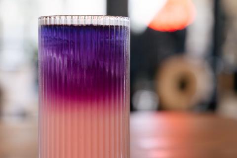 The galaxy lemonade sits in a tall fluted glass on a wooden table. It is light pink on the bottom and it blends into magenta and purple before reaching the blue pea flower tea at the top. It shimmers with edible glitter.