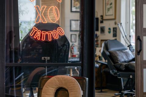 A wooden chair with a neon XO Coffee and Tattoo sign outside a tattoo studio with lots of natural light and enclosed by large window panes. 