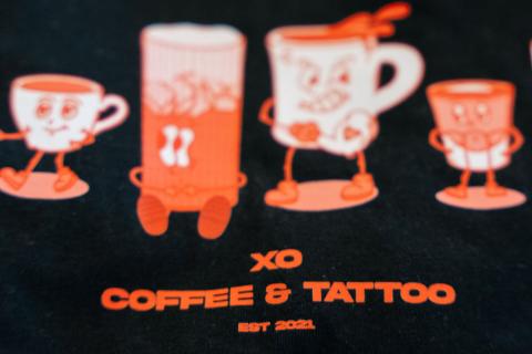 XO Coffee and Tattoo estimated 2021 is printed in red letters on a black T-shirt. Above the words are four animated characters - a teacup, a tall iced drink, a mug of coffee, and a shot of espresso.