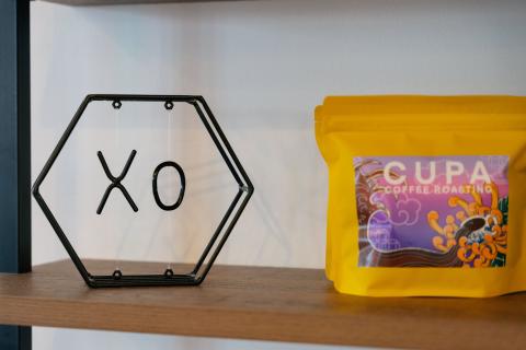 A hexagon-shaped black metal decoration with XO in the center next to a yellow bag of coffee grounds named Cupa.