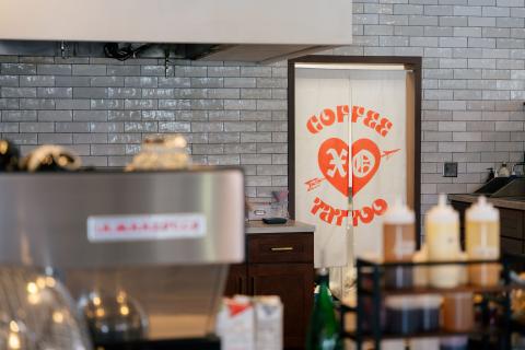 A shot of the interior of XO Coffee shows various coffee making tools and ingredients. In the background, there is a two paneled beige canvas curtain to the kitchen with XO Coffee and Tattoo in bright red letters.