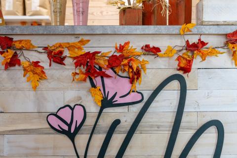 The counter at Madilynns Craft Coffee Shop is made of light wood planks and adorned with a cursive M and pink flowers. The back ground has the seasonal specials on a chalk board and the bagged coffee beans for sale.