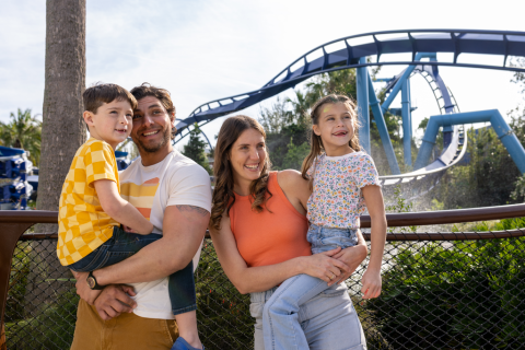 A family of four smiles and poses together in front of a looping roller coaster at a theme park, with parents holding their two young children.