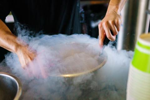 A person uses a ladle and tongs to prepare a dish in a bowl filled with swirling vapor, creating a dramatic cloud of smoke.