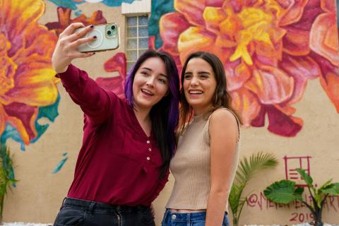 Two women take a selfie in front of a floral mural.