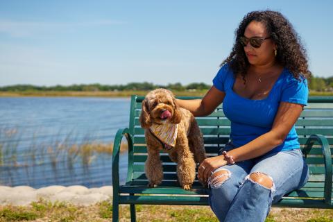 A woman sitting on a green bench by the lake in St. Cloud, Florida, petting her small brown dog who is wearing a colorful bandana.