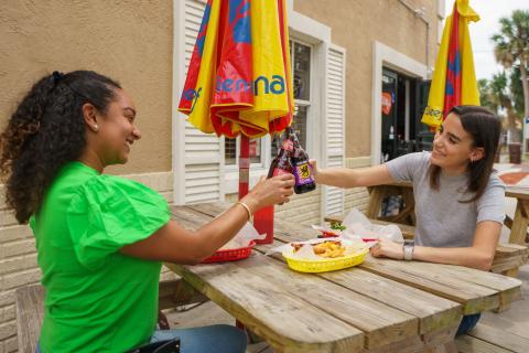 Two women toast with grape sodas at an outdoor picnic table, enjoying hot dogs and fries at Willy’s Wieners.