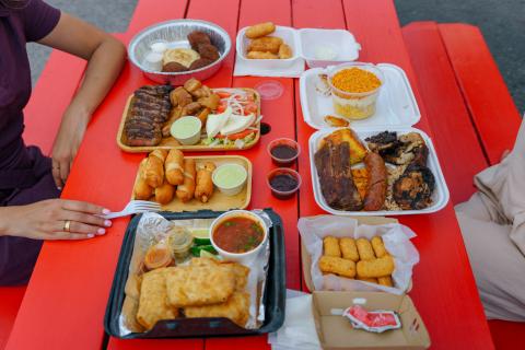 Overhead view of a colorful assortment of food truck dishes and drinks on a bright red picnic table, with Maryam Ishtiaq and Jen Bonner seated on either side, ready to enjoy the meal.
