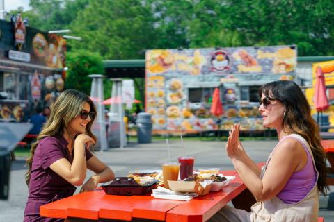 Maryam Ishtiaq and Jen Bonner sitting together at a bright red picnic table, laughing and enjoying a variety of food truck dishes and drinks at Food Trucks Heaven in Kissimmee.