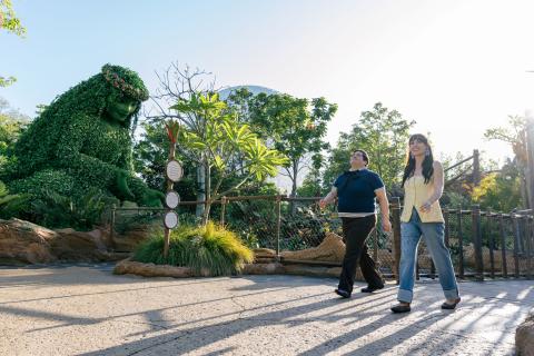 Harvey Guillén and Jen Bonner walk into Journey of Water at EPCOT®, with the sun rising and lush greenery — including Te Fiti — in the background.