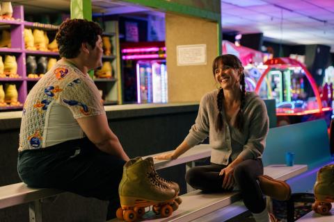 Harvey Guillén and Jen Bonner sit and chat at Skate Reflections, with skates in the foreground and arcade games behind them.