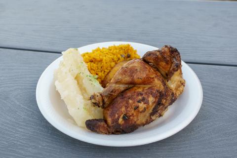 A takeout plate with rotisserie chicken, yellow rice with pigeon peas, and boiled yuca with garlic sauce, served on a white paper plate on a gray picnic table.