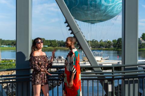 Sasheer Zamata and Jen Bonner chat and smile at Disney Springs®, with the Aerophile balloon visible in the background.