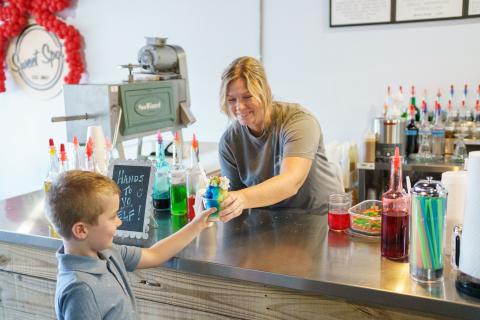 A smiling employee at Sweet Spot Snoballs in Downtown St. Cloud hands a colorful rainbow snoball to a young boy at the counter, surrounded by bottles of flavored syrups.