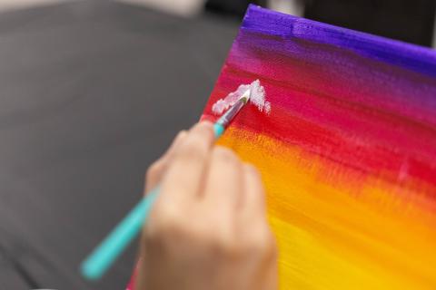 Close-up of a hand painting a white cloud on a vibrant pink, orange, and purple sunset canvas during a painting class.